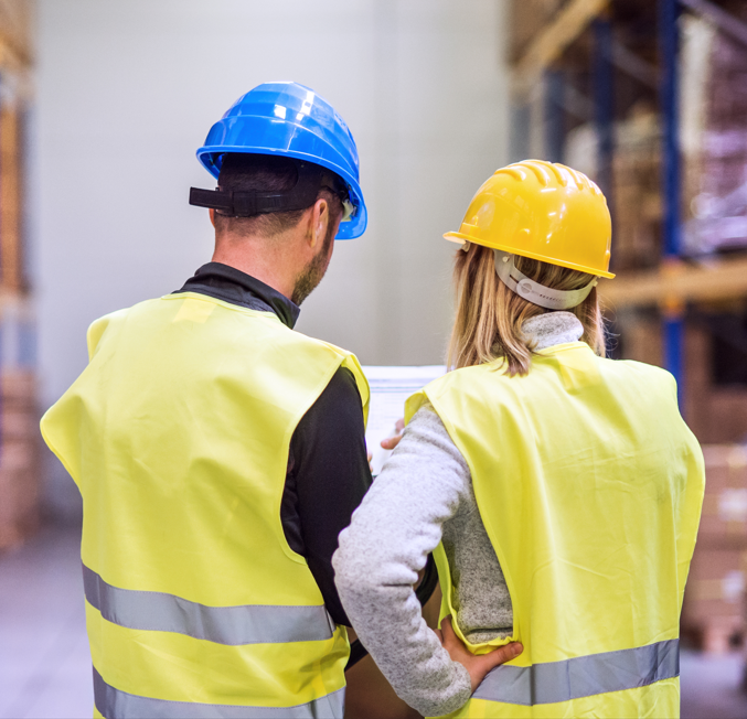 Two warehouse workers in safety vests and hard hats reviewing information inside a distribution facility